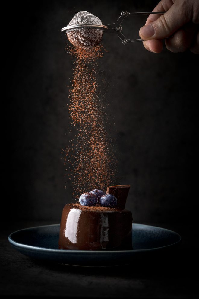 Cacao powder sprinkled on top of glazed chocolate cake with blueberries, photograph by Lauren Caris Short of Food Photography Academy