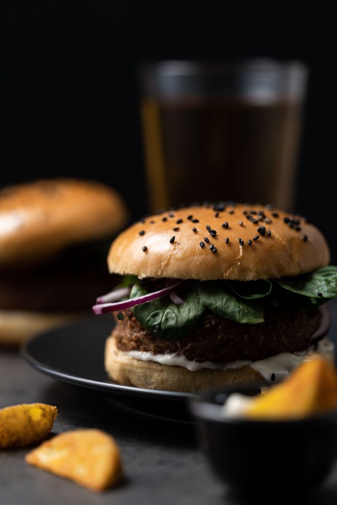 A cheeseburger, with a glass of beer, sauce cup, and chair in the background. Set against a black backdrop. Photograph by Lauren Caris Short of Food Photography Academy.