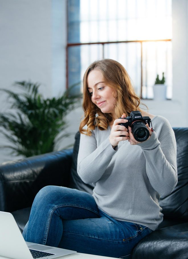 Professional food photographer, Lauren Short, sitting on a sofa holding her camera and looking at her laptop during the photo editing process. 