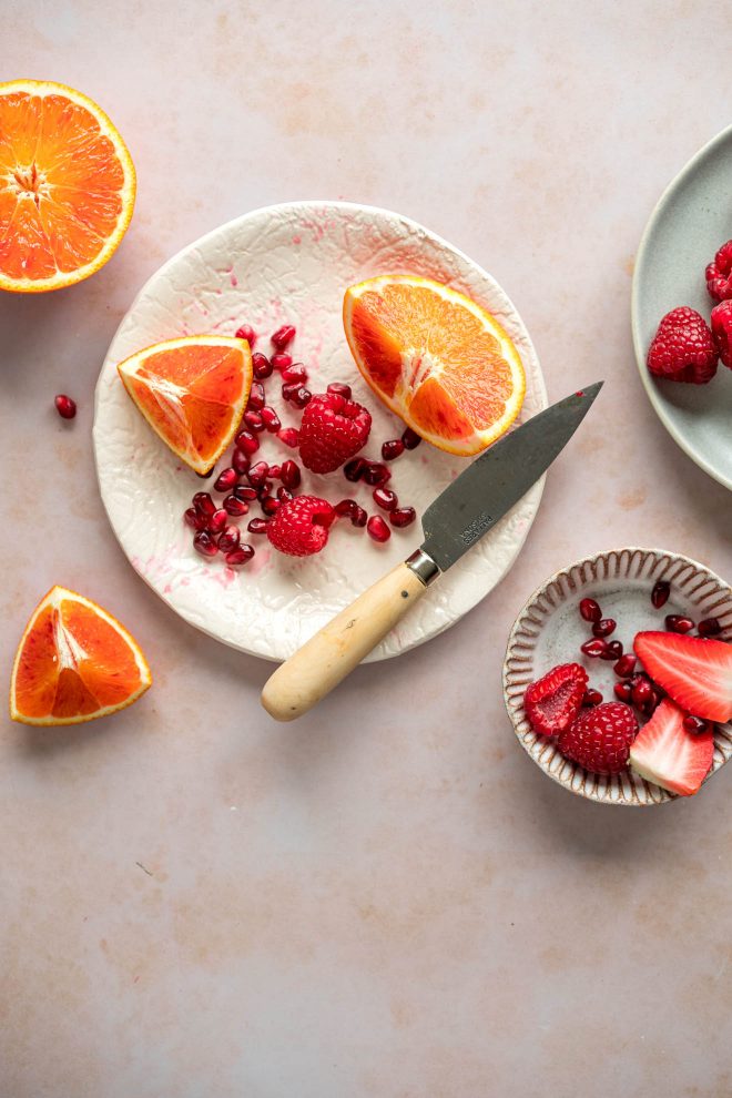 Orange slices, pomegranate seeds, raspberries, and strawberries with a knife on textured plates on a light, marbled surface. Photograph by Lauren Caris Short of Food Photography Academy.