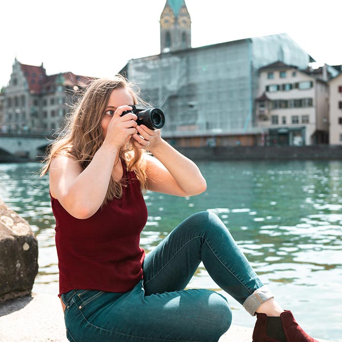 Lauren Caris Short sits near a river in the city with her camera to her eye