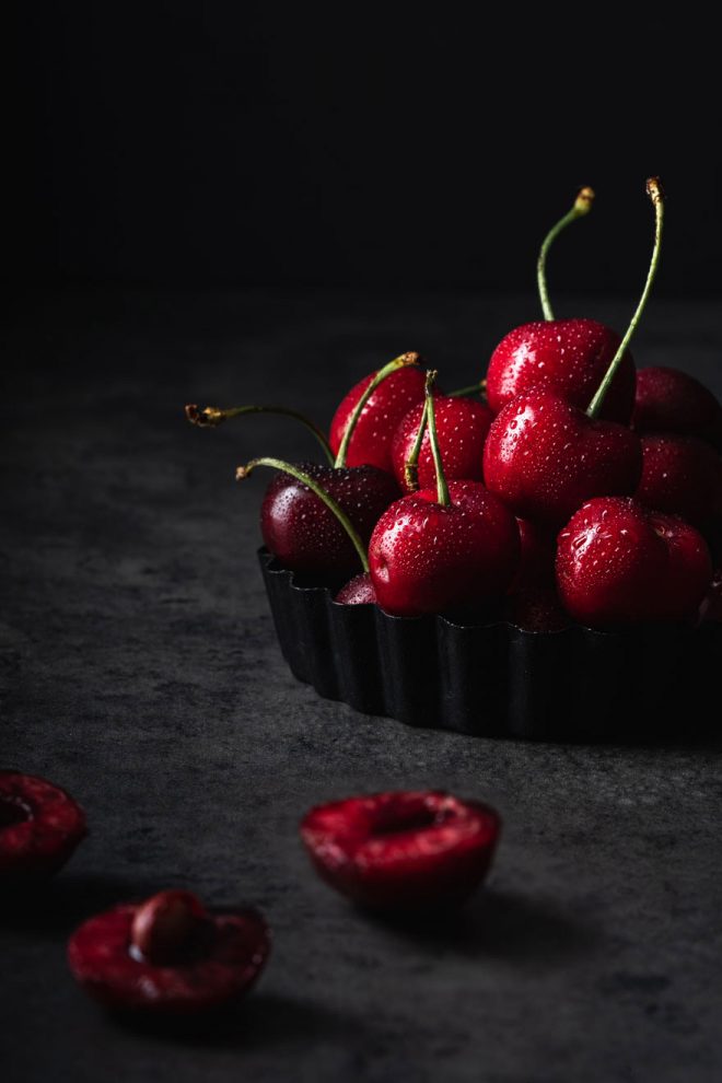 A small mountain of cherries rests in a tray with an easy dark and moody lighting setup by Lauren Short from Food Photography Academy.